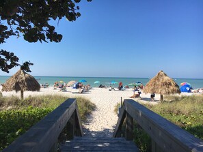 Ein Strand mit strohgedeckten Hütten, einer Holzpromenade, die zum Strand führt, und blauem Himmel.