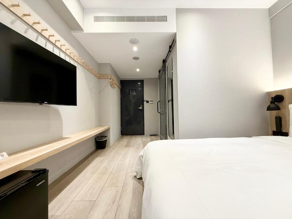 A modern hotel room with a large bed, a flat-screen TV mounted on the wall, a wooden bench, and a trash bin.