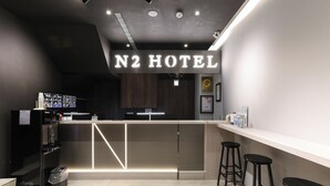 Zona de recepción moderna del hotel N2 con mostrador, taburetes de bar, refrigerador y un bote de basura.