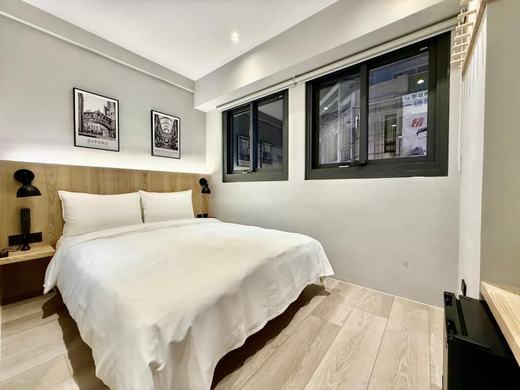Un dormitorio con cama, dos cuadros enmarcados, una mesita de noche, una lámpara y una ventana con vistas a un edificio.