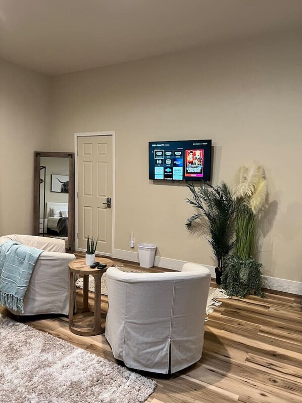 Una sala de estar con un televisor de pantalla plana montado en la pared, una puerta, una mesita con una planta y una planta grande en maceta.