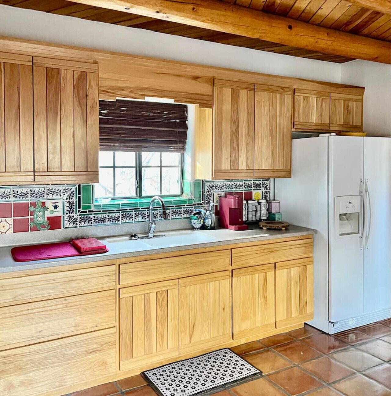 Une cuisine avec des meubles en bois, un réfrigérateur blanc, une fenêtre avec un store marron et un pare-feu en carrelage.