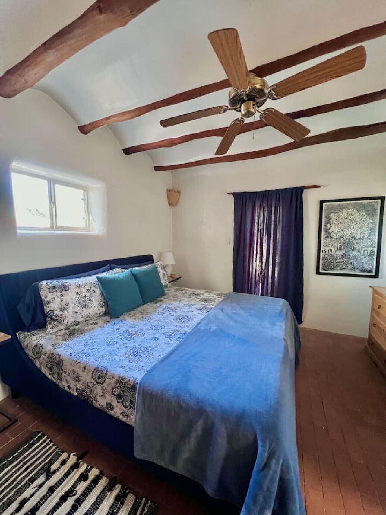 A bedroom with a bed, a ceiling fan, a window, a painting, and a wooden dresser.