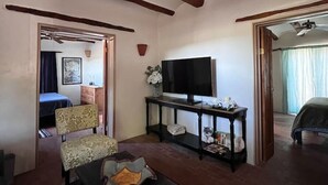 Un salon avec un ventilateur de plafond, un téléviseur posé sur un meuble, des fauteuils à motifs, une table basse et un tapis rouge.