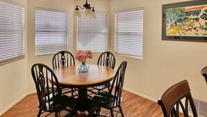 A dining room with a round wooden table, six black chairs, and a vase with pink flowers. There are three windows with blinds, a ceiling fan, and a painting on the wall. - 2 bd/2bth near NMSU and Memorial Medical Center (Las Cruces)