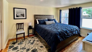 Un dormitorio con cama, una silla, una mesita de noche, una lámpara y una ventana con cortinas.