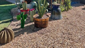 Un patio con una vela azul de sombra, varias macetas con plantas y una cerca de madera.