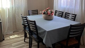 A dining room with a round table covered with a light blue tablecloth, surrounded by six wooden chairs. A vase with pink flowers is placed in the center of the table. The room has beige walls and a wooden floor. - 1 bed W/D in unit, soaker tub near Field of Dreams (Las Cruces)