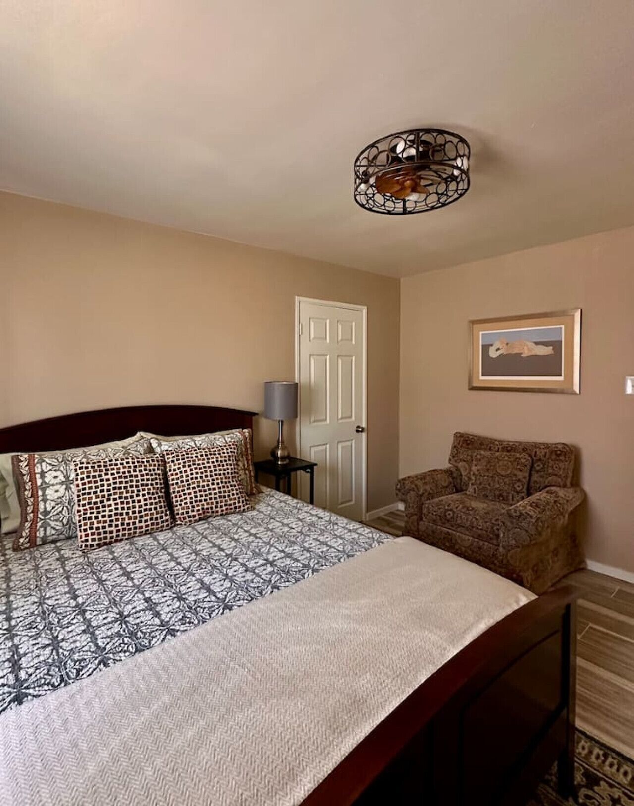 Een slaapkamer met een bed, een stoel, een lamp, een deur en een lijst met een afbeelding aan de muur.