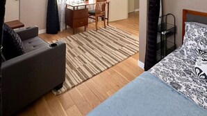 A compact room with a bed, a sofa, a dining table with chairs, and an air conditioning unit.