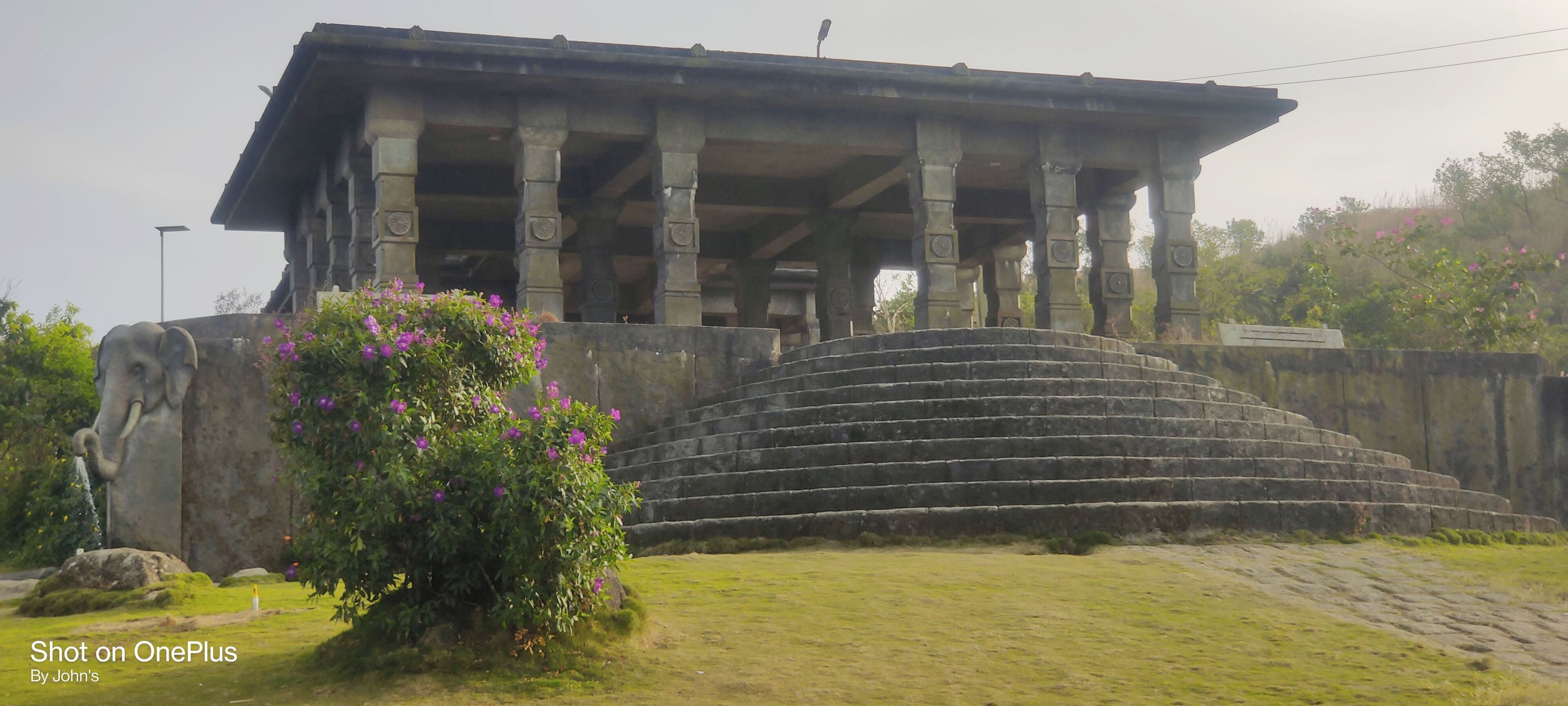 A large stone structure with a roof supported by columns, a staircase leading up to it, and a statue of an elephant.