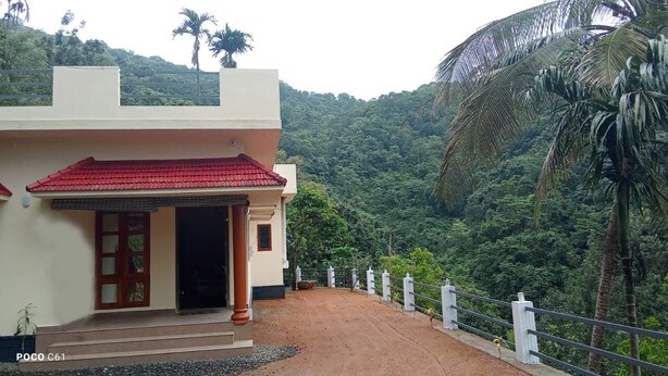 A building with a red-tiled roof, a white facade, and a red door. It is situated on a dirt path with a white fence and palm trees in the background.