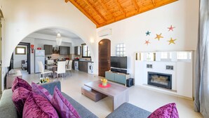A spacious living area with a wooden ceiling, a fireplace, a TV, and a kitchen in the background.