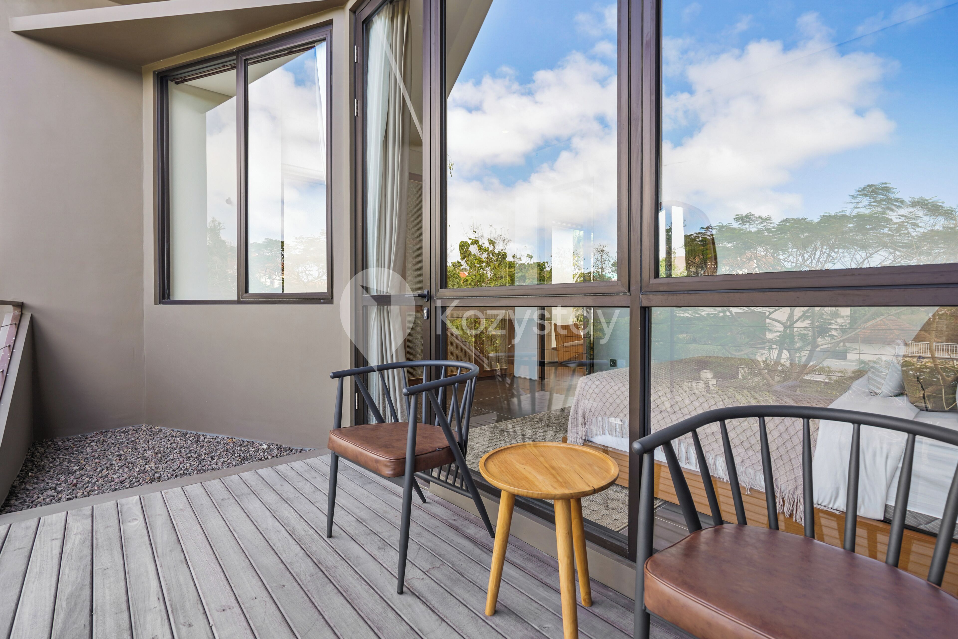 Ein Balkon mit Holzboden, zwei Stühlen, einem kleinen runden Tisch und einem großen Fenster mit Blick auf Gebäude und Bäume.