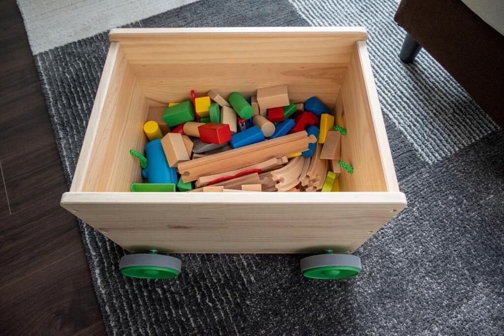 Eine hölzerne Aufbewahrungsbox, gefüllt mit bunten Holzblöcken und -zylindern auf einem Teppichboden.
