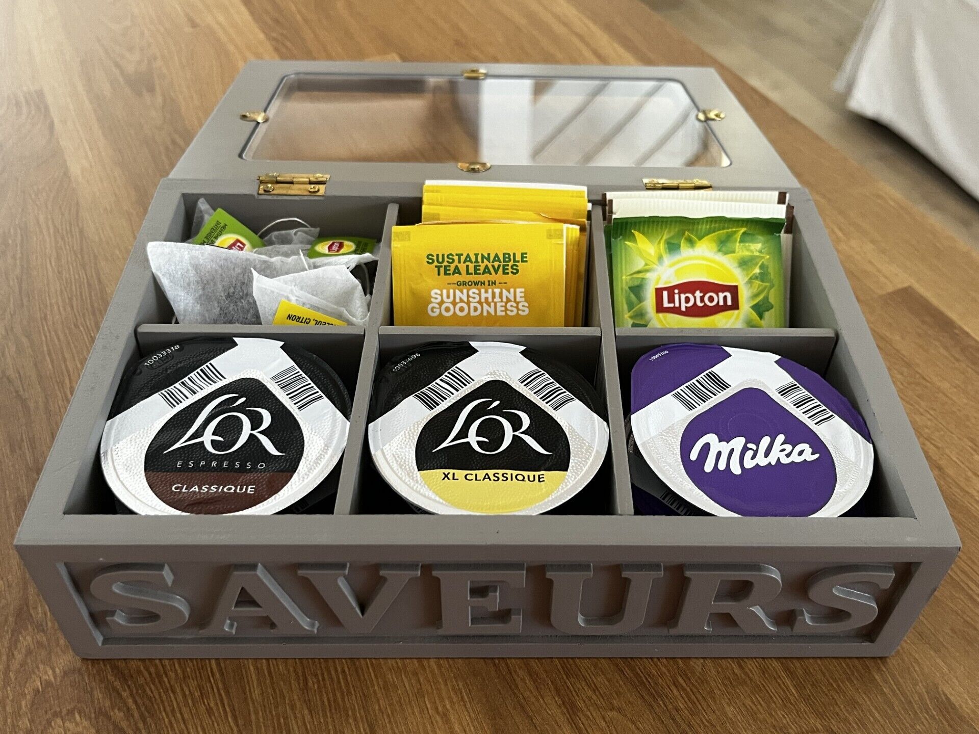 Una caja de almacenamiento gris con la etiqueta 'SAVEURS' y compartimentos que contienen bolsitas de té y un paquete de té Lipton.