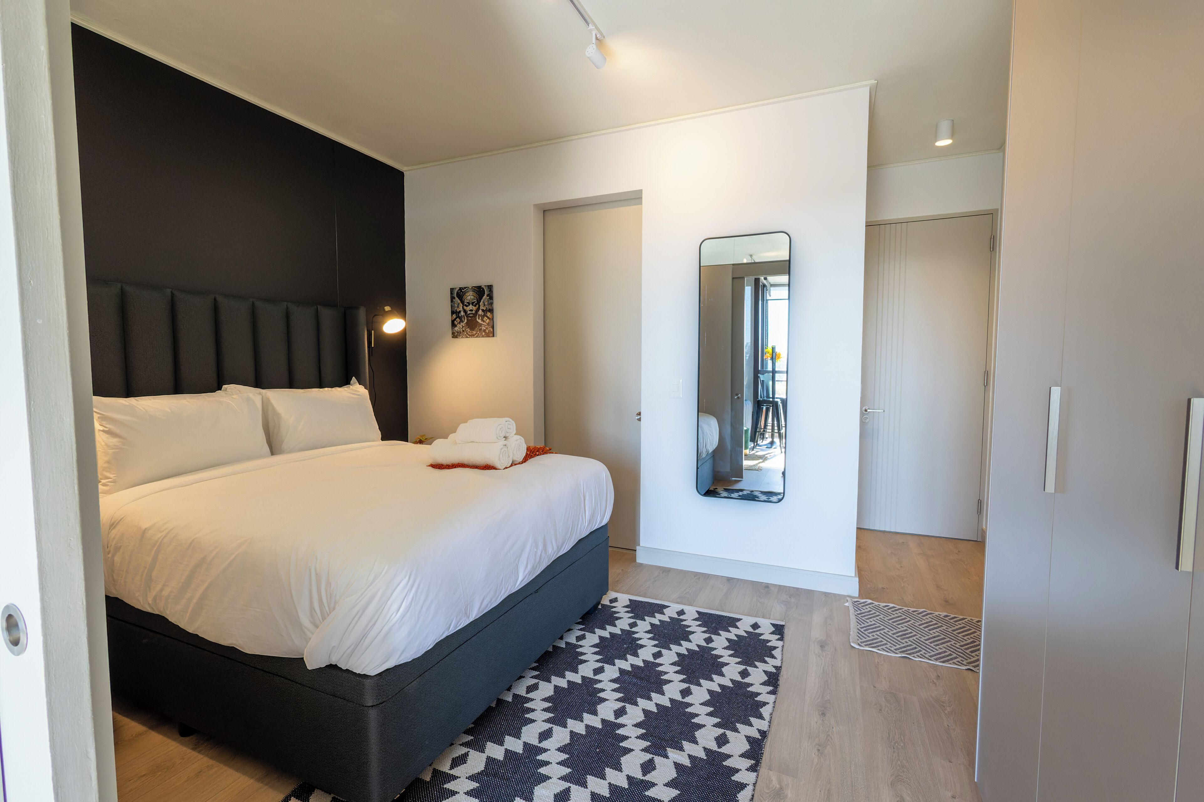 A modern bedroom with a large bed, a dark headboard, a patterned rug, and a mirror on the wall.