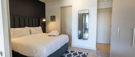 A modern bedroom with a large bed, a dark headboard, a patterned rug, and a mirror on the wall.