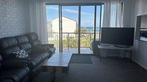Ett vardagsrum med en svart lädersoffa, ett soffbord, en tv på ett tv-bord och en balkong med utsikt över havet och byggnaderna.