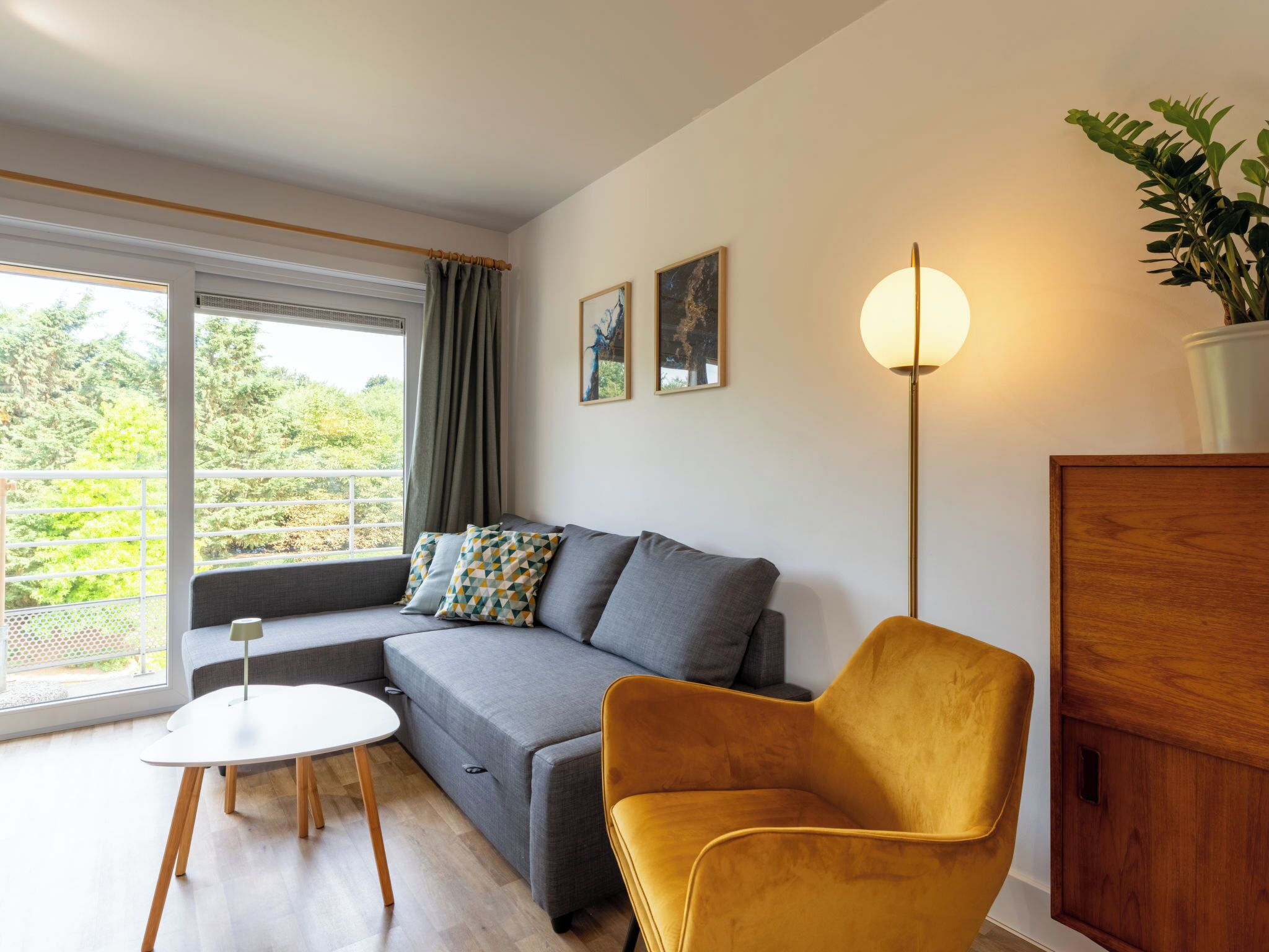 Un salon moderne avec un canapé gris, un fauteuil jaune, une table blanche et un lampadaire. On y trouve une fenêtre avec des rideaux, un tableau au mur et une plante en pot.