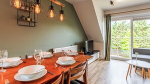 A dining area with a wooden table set for four, a television on a stand, and a large window with a view of greenery.