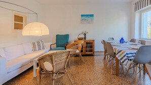 A cozy room with a white sofa, wicker chairs, a dining table with striped tablecloth, and a painting of a beach scene.