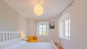 A bedroom with a large bed, a small table with a lamp, a yellow chair, a window with curtains, and a picture on the wall.