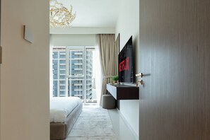A modern bedroom with a large bed, a TV mounted on the wall, and a view of high-rise buildings through the window.