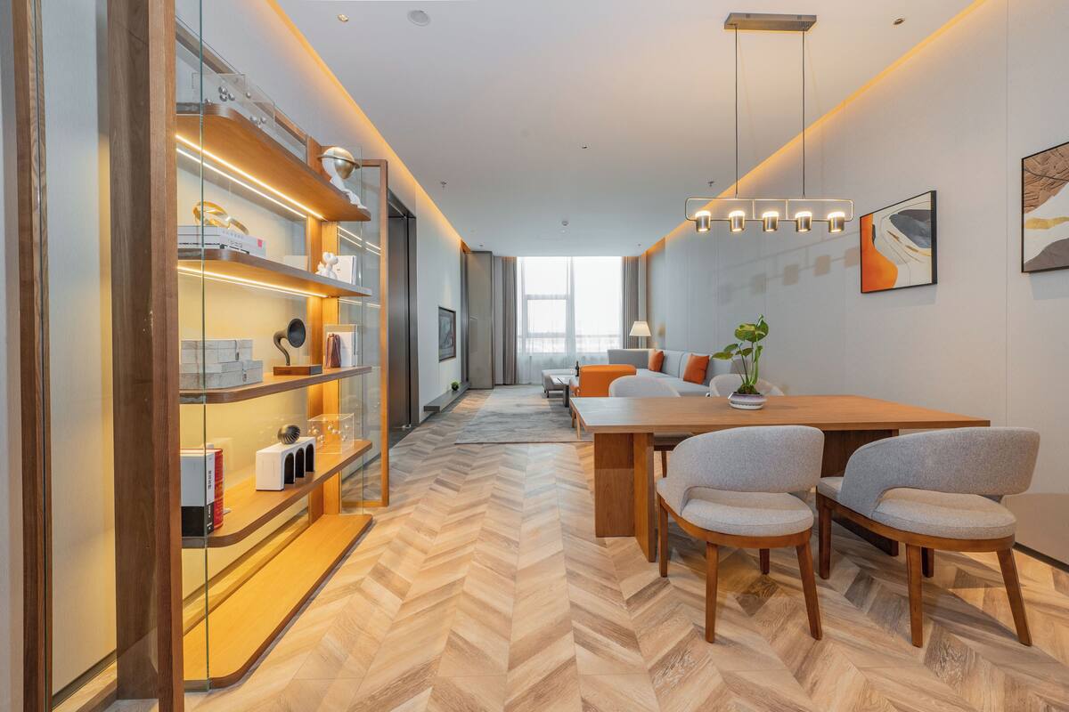 Ein modernes Hotelzimmer mit Holzdielen in Fischgrätmuster, einem Essbereich mit Holztisch und -stühlen sowie einer Glastrennwand mit Regalen für Bücher und Dekorationsobjekte.