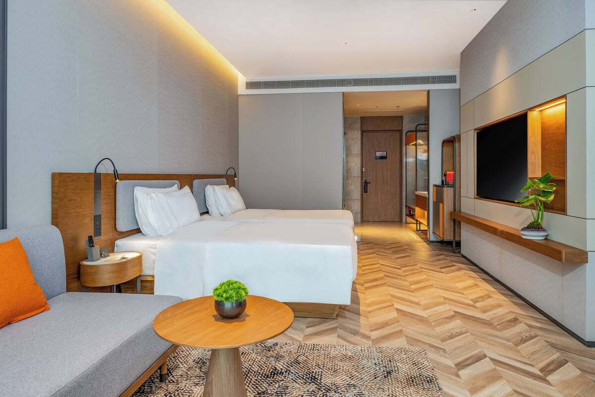 Ein modernes Hotelzimmer mit einem großen Bett, einer Couch, einem kleinen Tisch mit Pflanze, einem Fernseher und Holzboden.