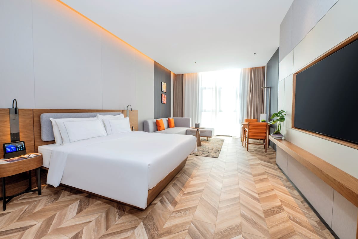 Ein modernes Hotelzimmer mit einem großen Bett, einem Schreibtisch mit Computer, einer Sitzecke mit Sessel und kleinem Tisch sowie einem großen Fernseher an der Wand.