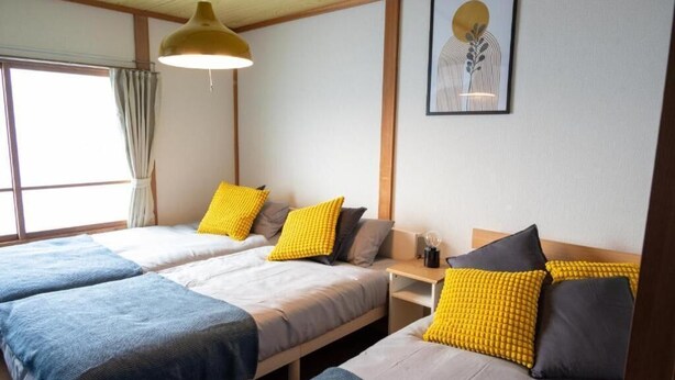 Une chambre avec deux lits, chacun recouvert d’une couvre-lit bleue et grise, et agrémentée d’oreillers jaunes. Un tableau encadré orne le mur, et une fenêtre est dotée de rideaux.