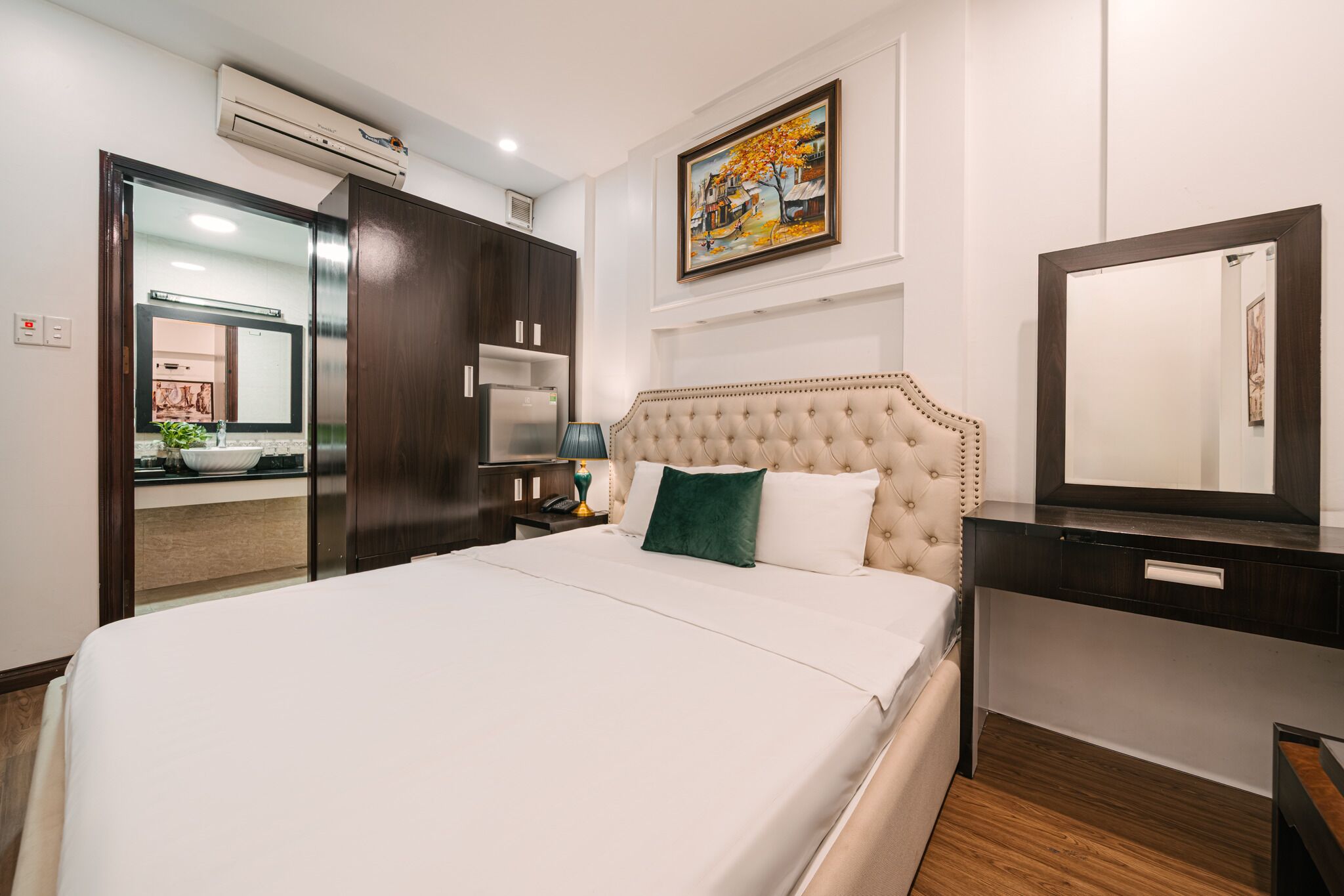 A hotel room with a bed, a dresser, a mirror, a painting, and an air conditioning unit.