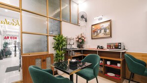 Een restaurant met een glazen scheiding, groene stoelen, een ventilator, een schilderij en een koffieapparaat.