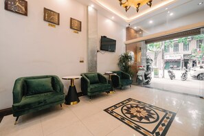 Un hall d’entrée avec des fauteuils à revêtement vert, une petite table ronde, un tapis décoratif et un téléviseur fixé au mur.