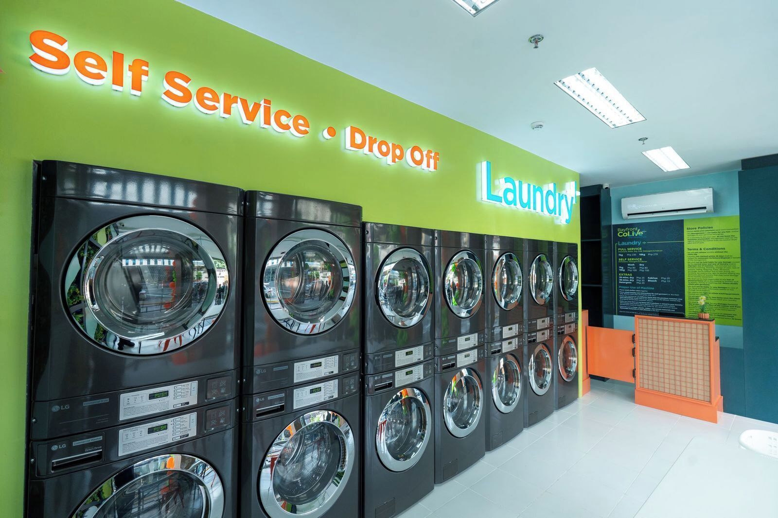 A self-service laundry area with multiple washing machines and dryers, a sign indicating self-service and drop-off options, and a Bayfront ColiVe store policy sign.
