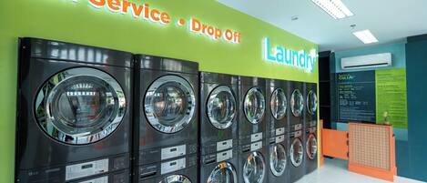 A self-service laundry area with multiple washing machines and dryers, a sign indicating self-service and drop-off options, and a Bayfront ColiVe store policy sign.
