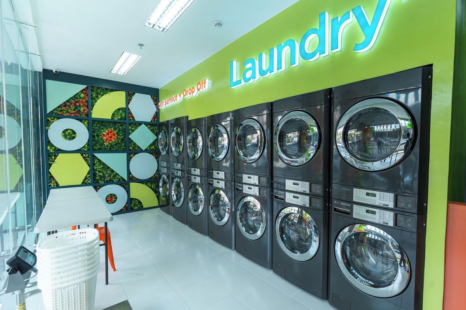A self-service laundry area with multiple washing machines, a bright green wall with the word 'Laundry' and a sign indicating self-service drop-off.
