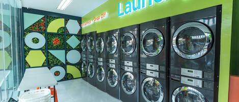 Zona de lavandería autoservicio con varias lavadoras, una pared verde brillante con la palabra 'Laundry' y un cartel que indica servicio de entrega y recogida.