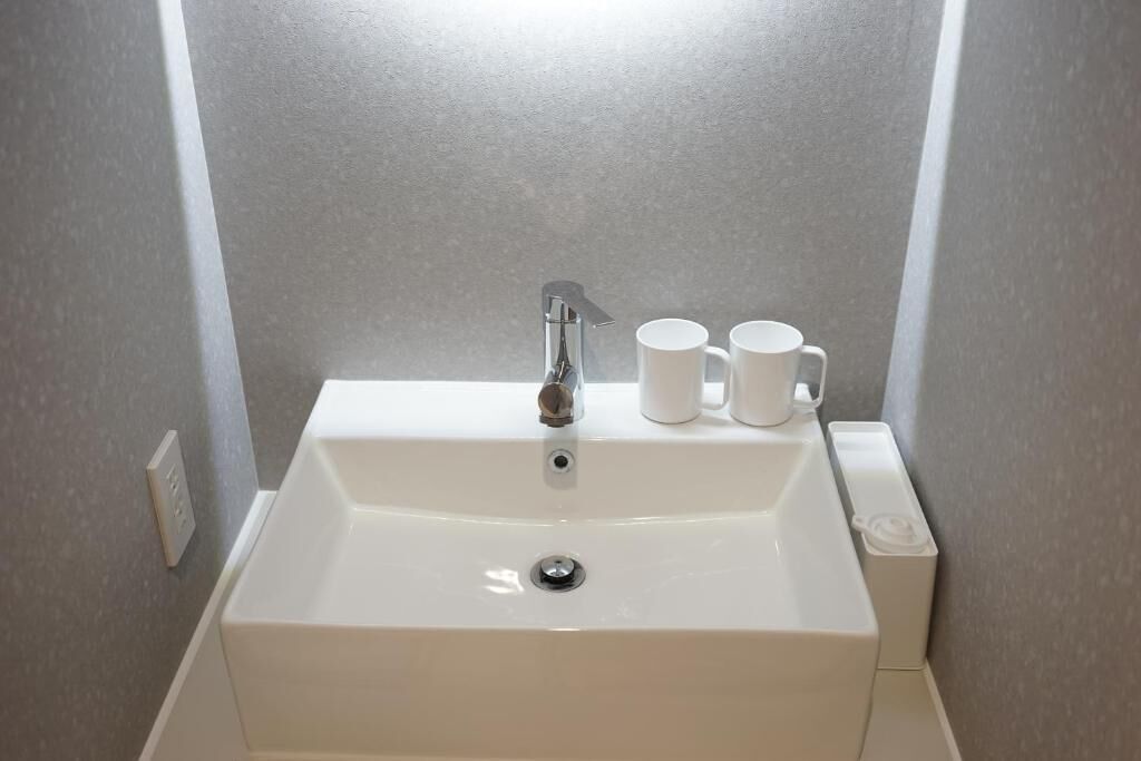 Un lavamanos blanco con un grifo moderno, dos tazas blancas y un pequeño recipiente blanco a la derecha.