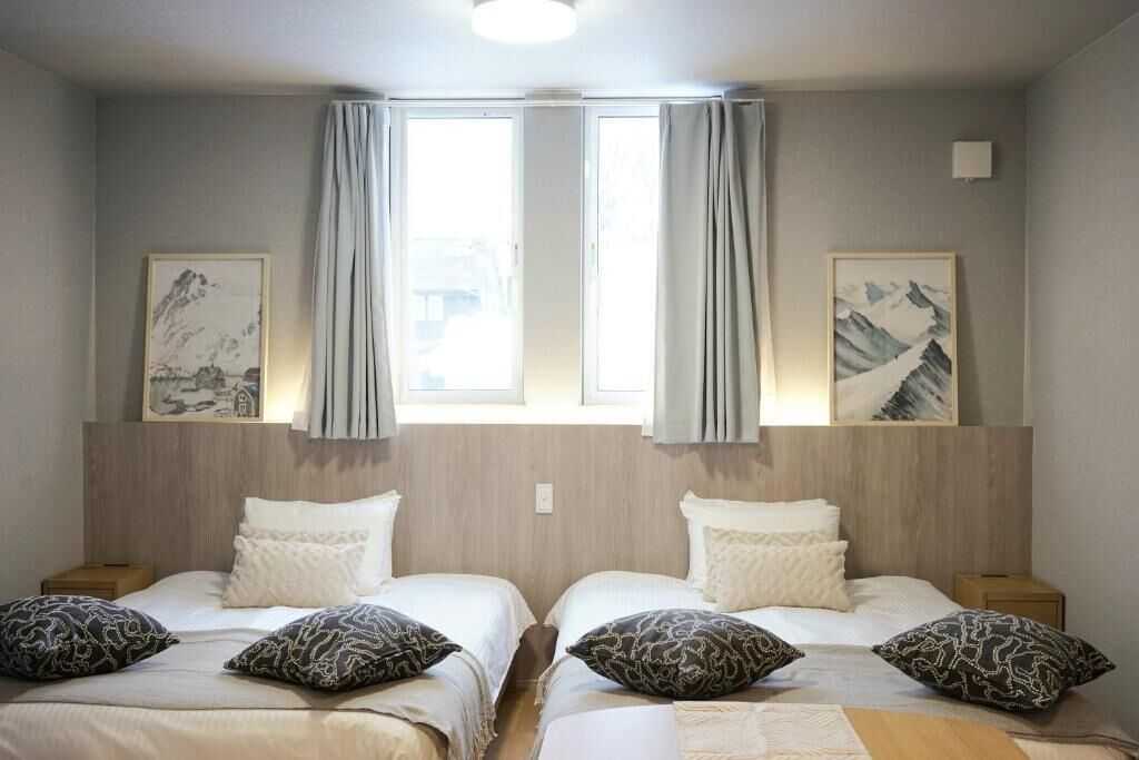 A hotel room with two beds, each with white bedding and patterned pillows. There are two framed pictures on the wall, a window with curtains, and a light fixture above the beds.