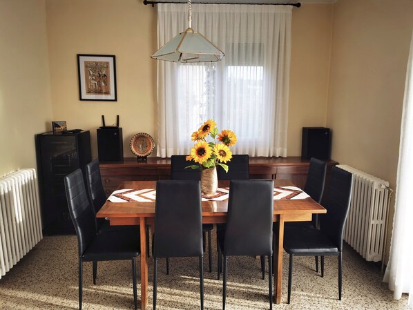 Un coin repas avec une table en bois, des chaises noires, un vase avec des tournesols, un tableau encadré et un radiateur.