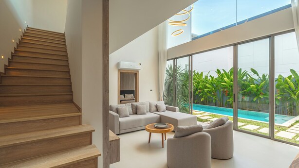 A modern interior with a staircase, a living area with a sofa and armchairs, a coffee table, and a view of a swimming pool through large windows. - Near Beach & Famous Boat Ave V217 (Choeng Thale)