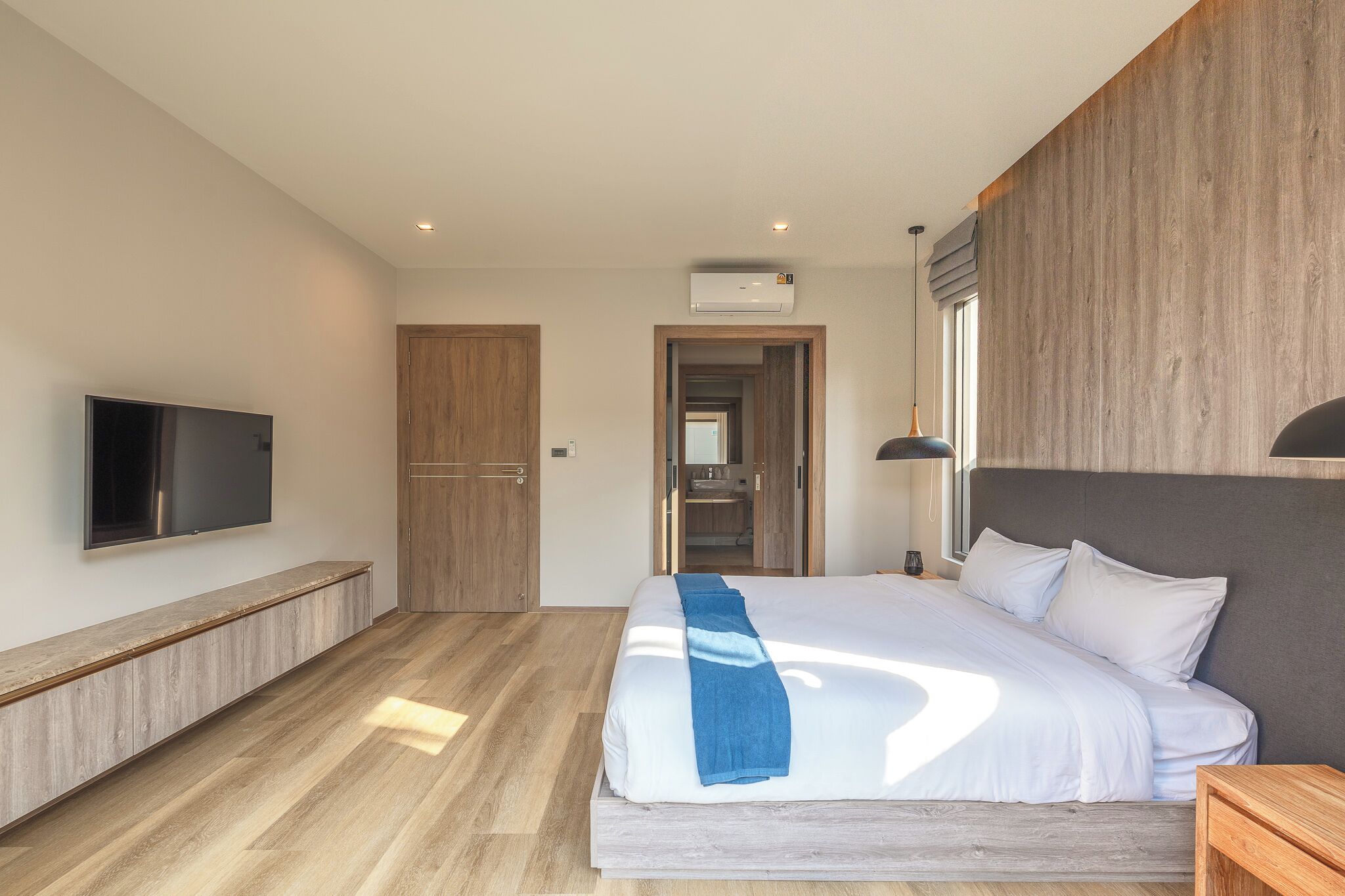 A modern hotel room with a large bed, a flat-screen TV, a wooden bench, and a view of the bathroom.