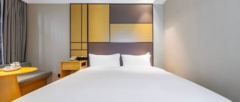 A modern hotel room with a large bed, a bedside table with a lamp, a small desk, and a chair.