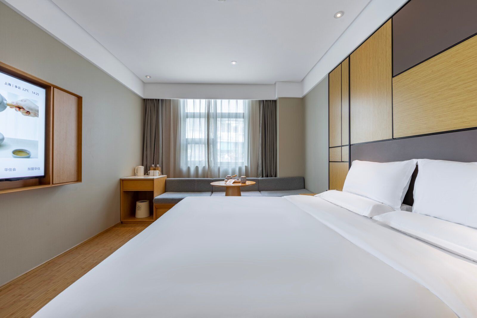 A modern hotel room with a large bed, a small seating area, a TV mounted on the wall, and a window with curtains.