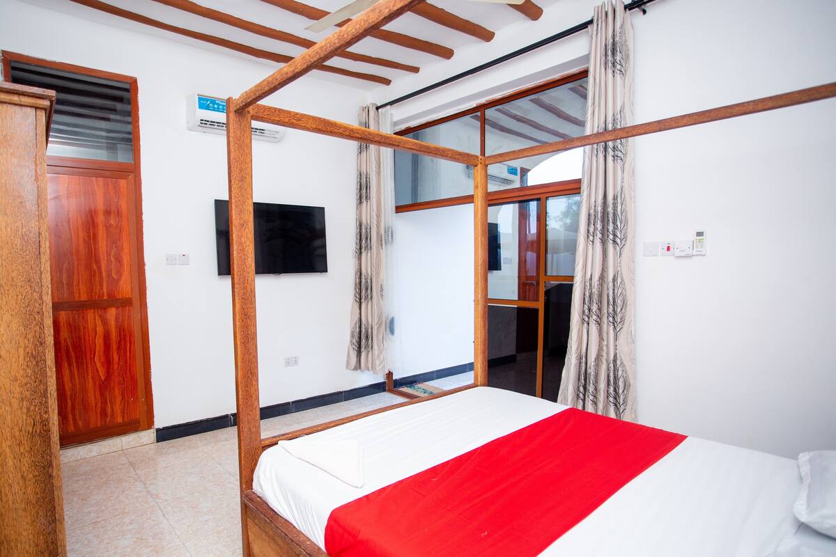 Una habitación con cama con dosel, puerta de madera, televisor montado en la pared y ventana con cortinas.