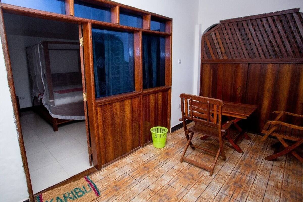 Una habitación con suelo de madera, una cama con mosquitero, una mesa de madera con sillas y una cesta verde.
