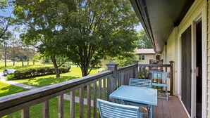 Una terraza de madera con muebles metálicos azules, vista a un césped cuidado y árboles, y una casa al fondo.