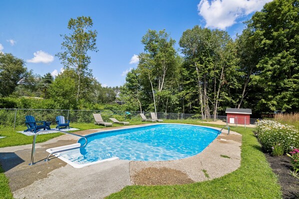Una piscina con solárium de hormigón, rodeada por una valla, y dos sillas Adirondack azules.